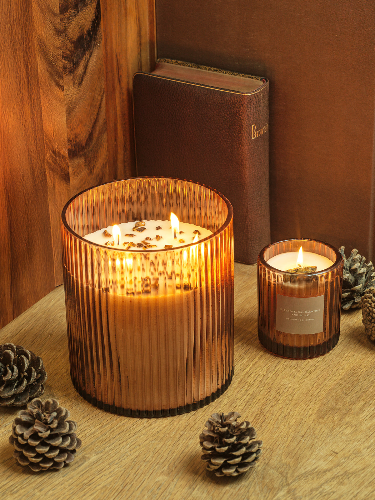 Two lit soy wax candles in ribbed glass holders containing tiger's eye on a wooden surface with pinecones.