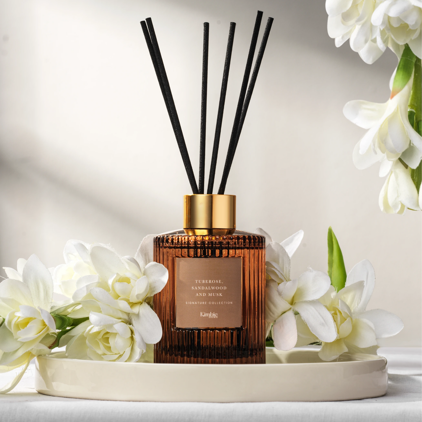 A natural oil diffuser with black sticks on a white tray surrounded by white flowers on a light background