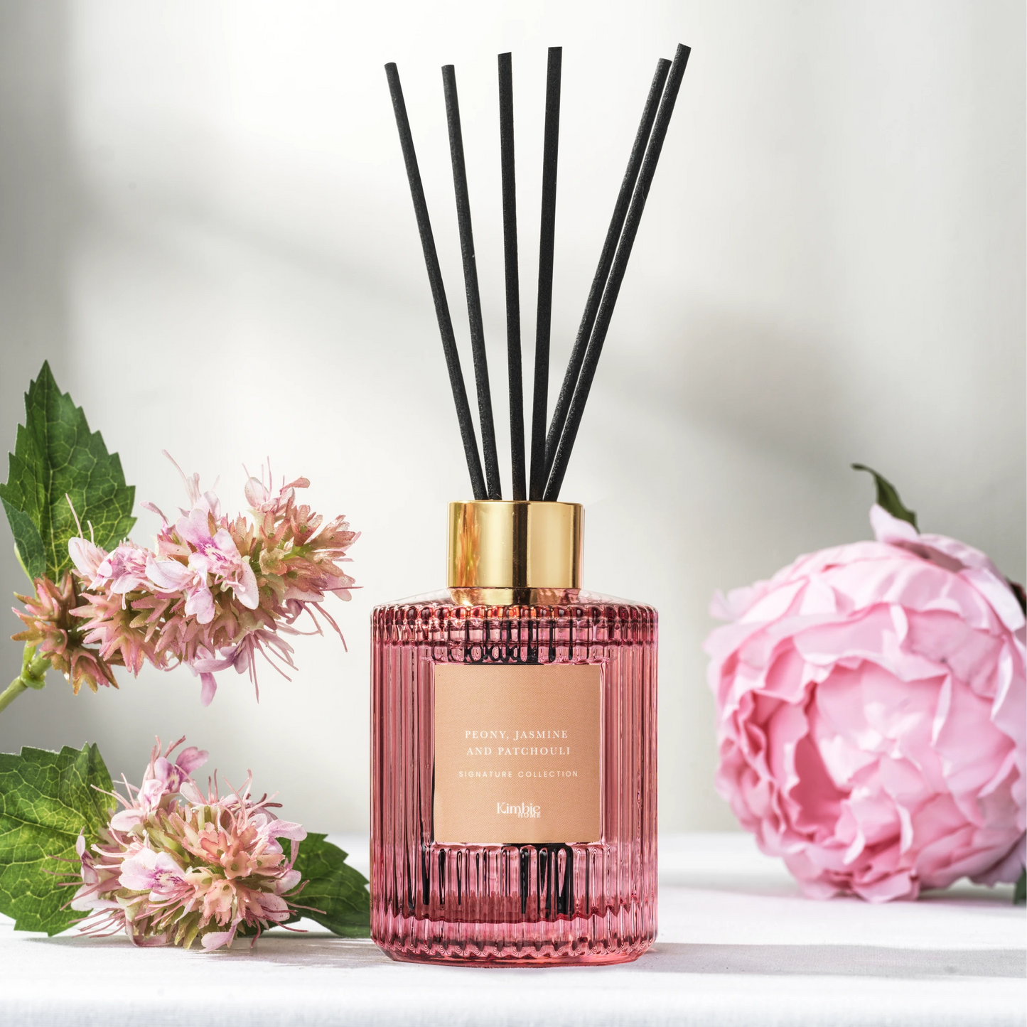 Pink natural oil diffuser bottle with black sticks surrounded by pink flowers and green leaves on a light background